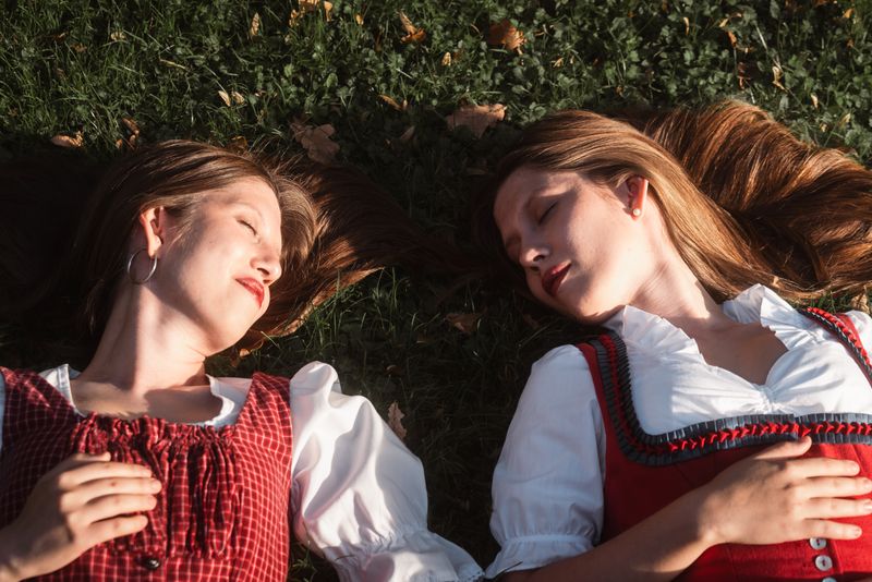 Twin sisters lie on grass wearing traditional dirndl dresses in Graz, Austria.