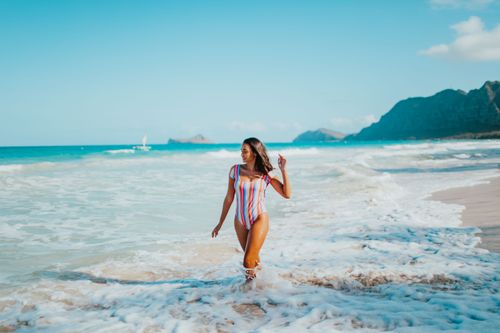 Free A woman in a colorful swimsuit walks on a sandy beach with waves and mountains in the background. Stock Photo