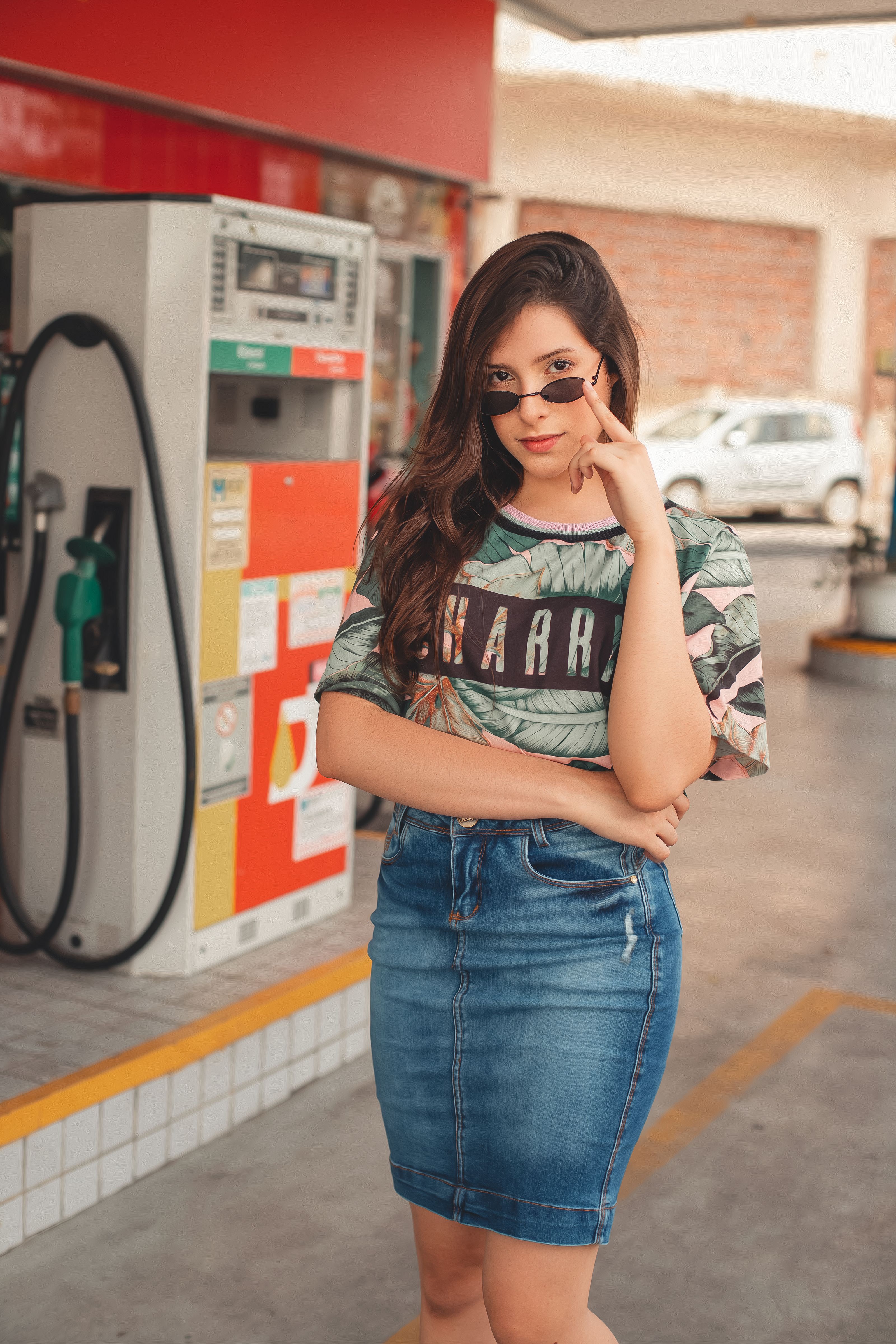 Mulher confiante em uma saia jeans e óculos de sol posando em um posto de gasolina, exibindo moda urbana.
