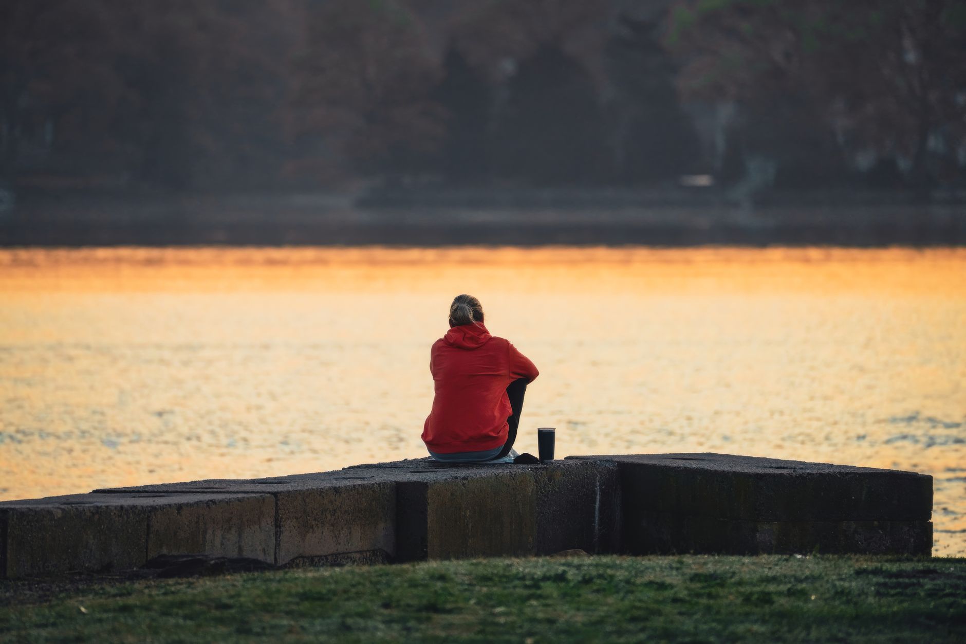 Person in red hoodie sitting by a lake at sunset, reflecting in serene solitude.