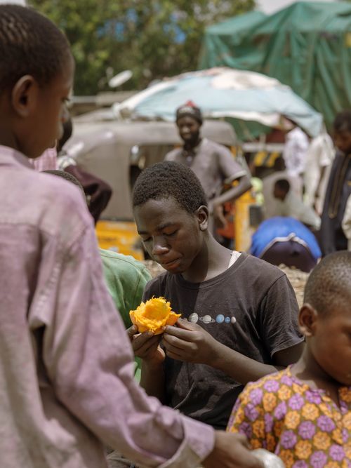 Free A young boy enjoys fruit in a bustling market in Zaria, Nigeria. Lively street atmosphere. Stock Photo