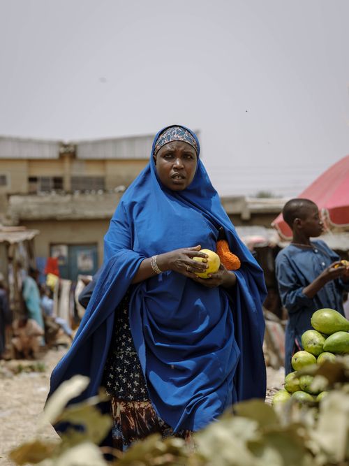 Free Vibrant street scene of an African woman in blue attire holding fruit at a market in Zaria, Nigeria. Stock Photo
