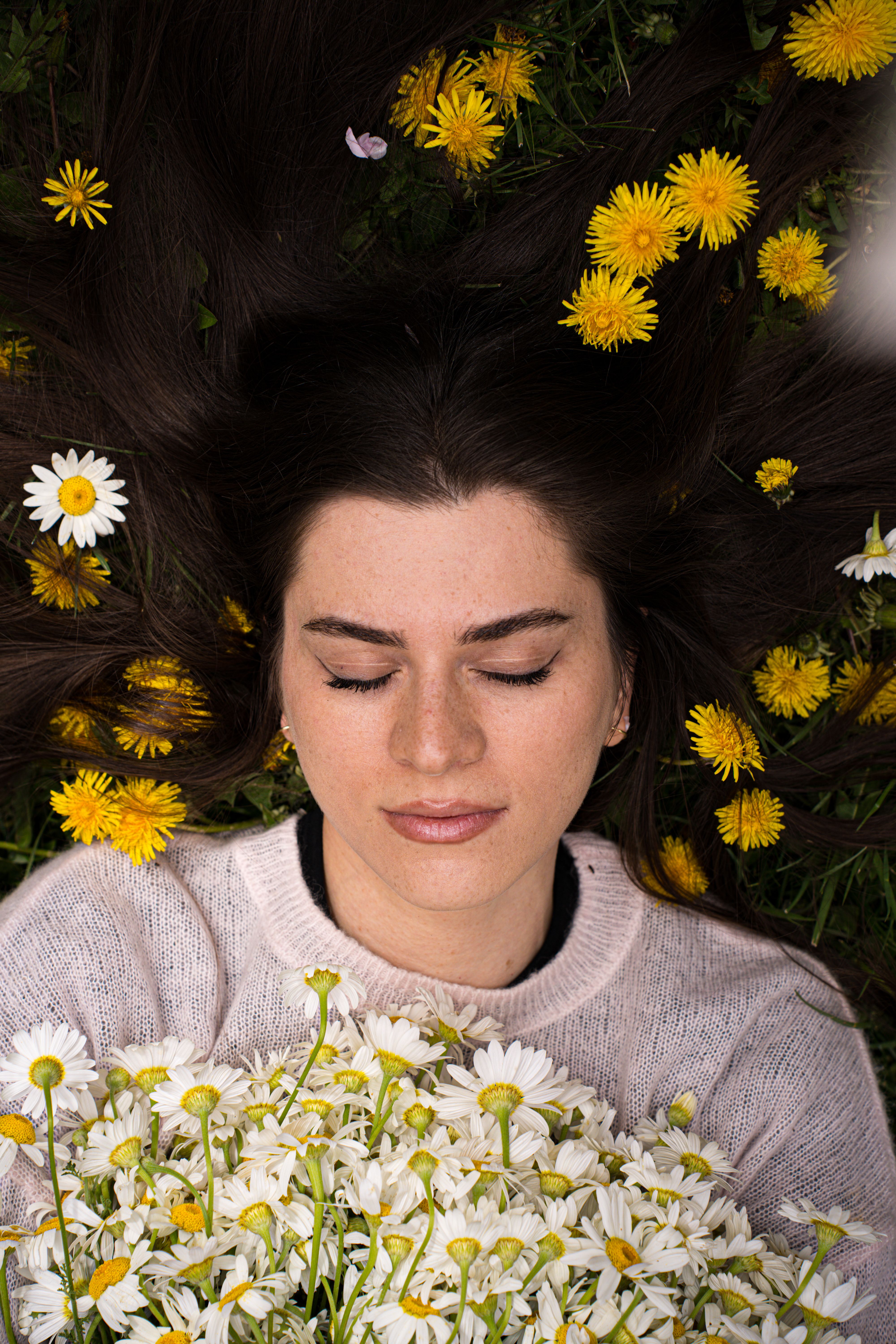 Woman lying among yellow and white daisies, eyes closed, relaxed expression, nature setting.