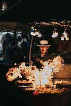 Night market vendor cooks street food on open flame with lights above, Xuan Huong, Da Lat.