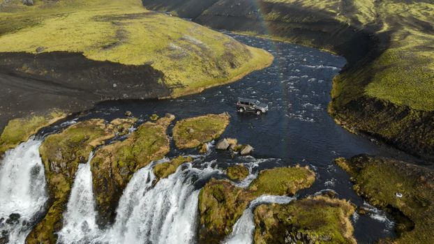 An off-road SUV navigates a river surrounded by lush Icelandic landscapes and waterfalls.