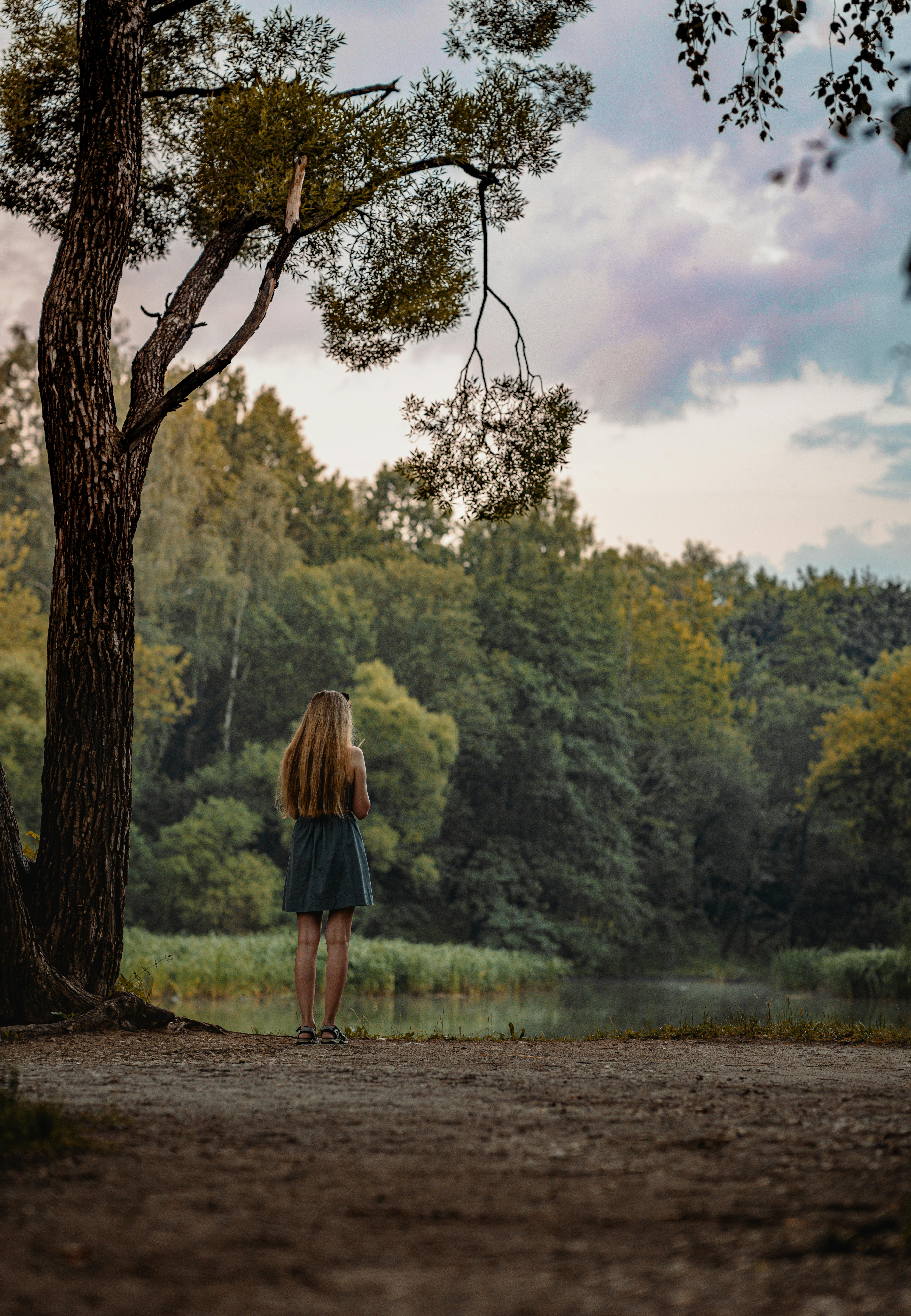 A young woman stands by a forest clearing, gazing at the lush greenery under a soft sky.