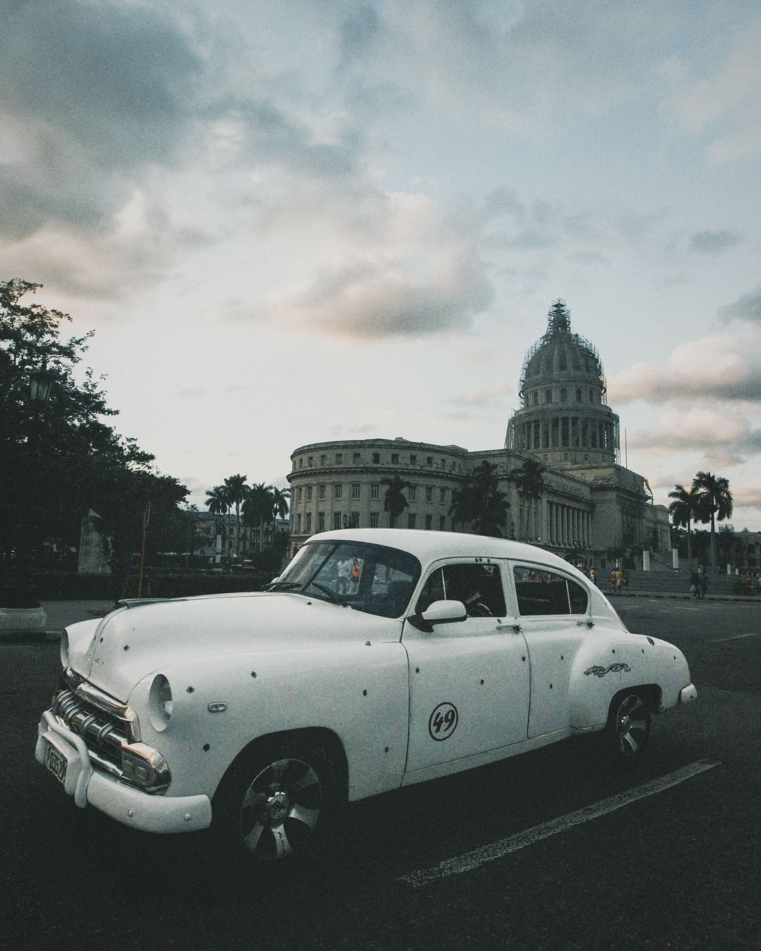 Vintage white car parked in front of a historic capitol building during the day.