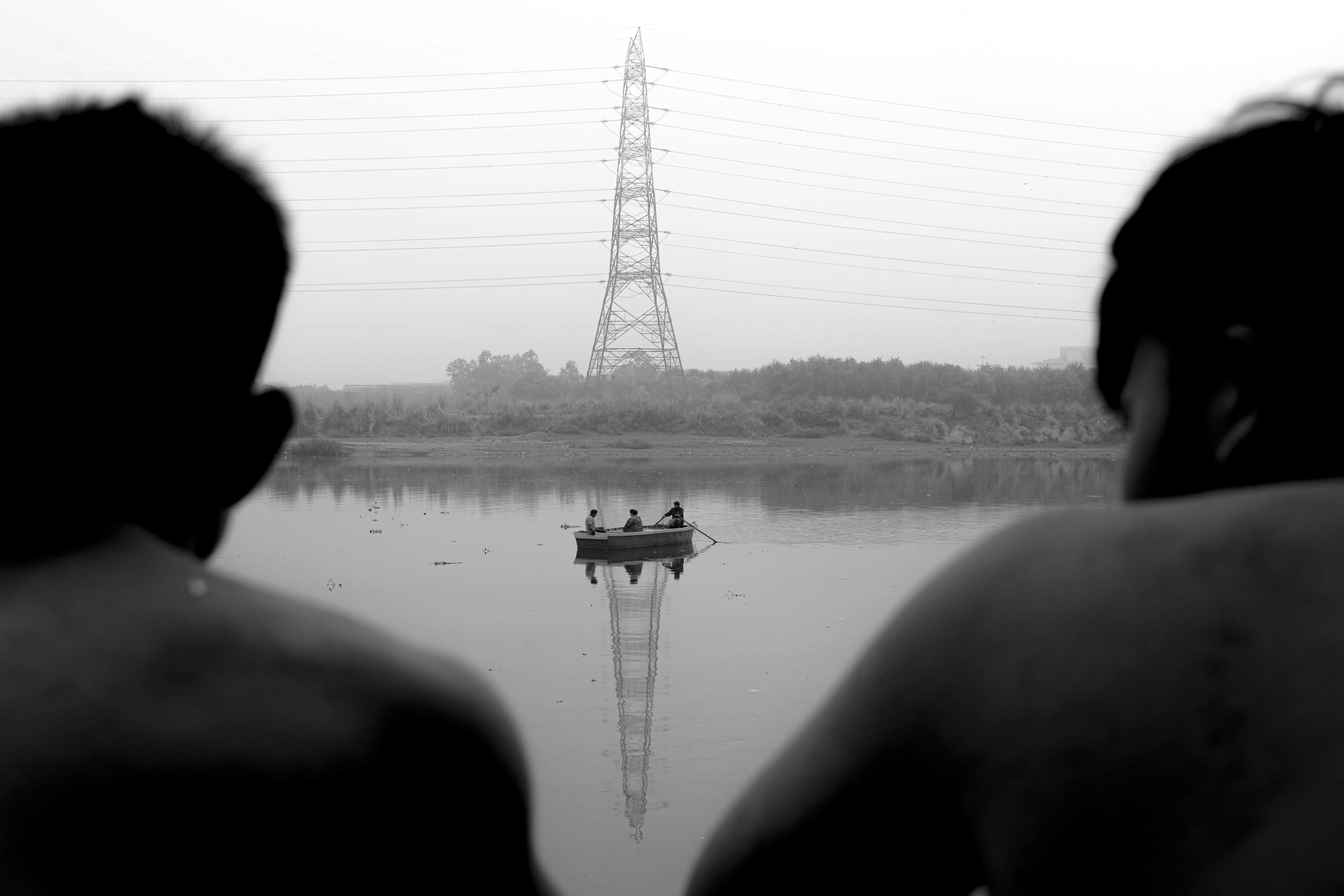 Silhouetted people watch a boat drift on a calm river with a distant powerline view.