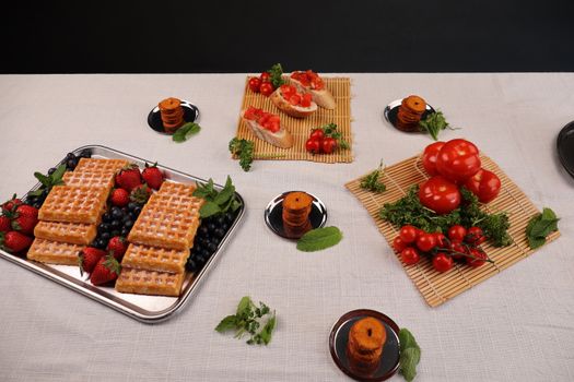 Aerial view of a brunch setup with waffles, berries, tomatoes, and bruschetta on a table.