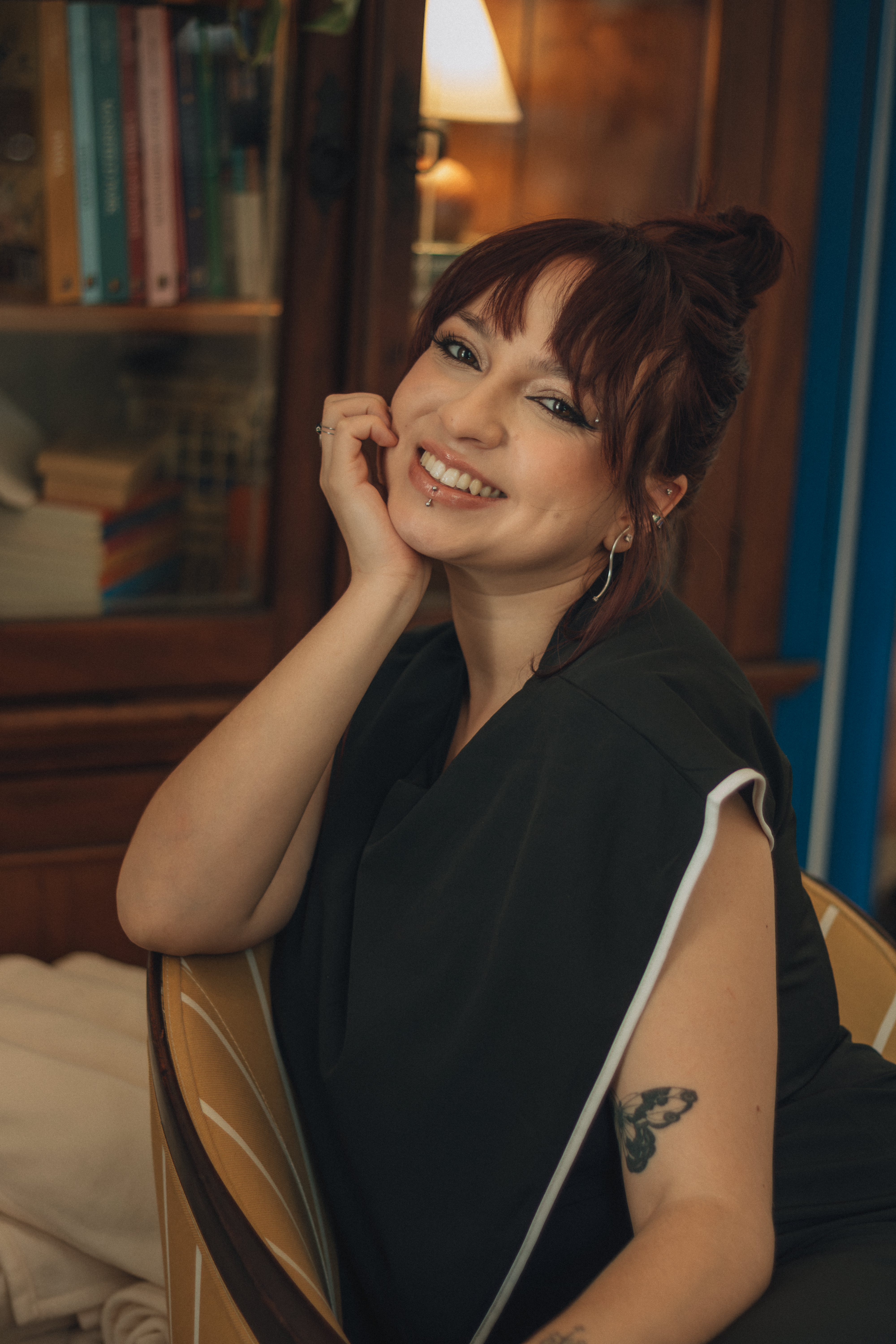 Cheerful woman in a cozy room, smiling and leaning on a chair, showing off her arm tattoo.