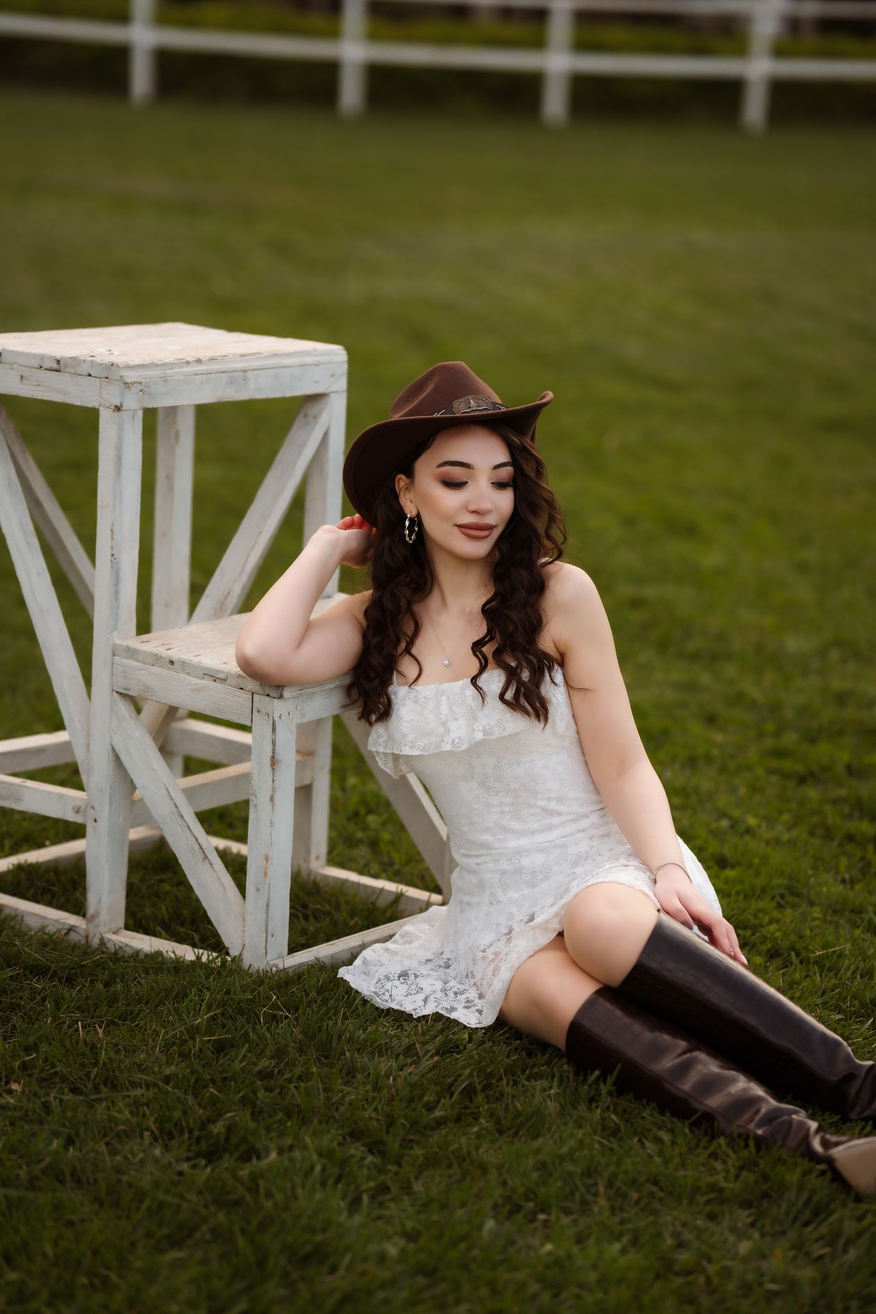 Woman in a white lace mini dress with tall brown boots and a brown hat sitting on grass