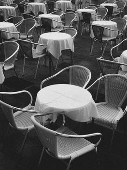 A black and white photo of a Venetian cafe setup with empty wicker chairs and tables covered in tablecloths.