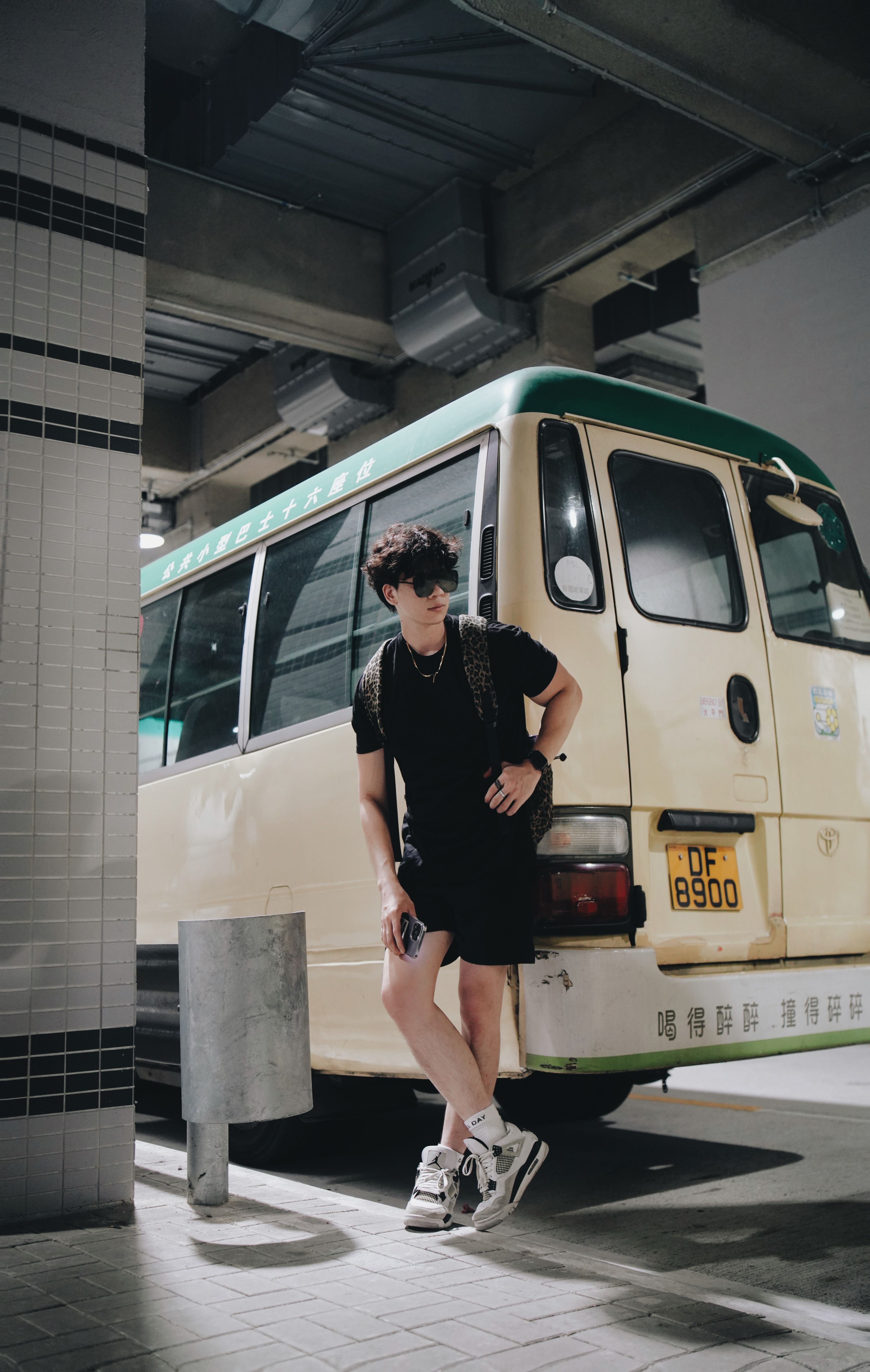 Young adult in urban outfit leans on a retro cream and green minibus indoors.