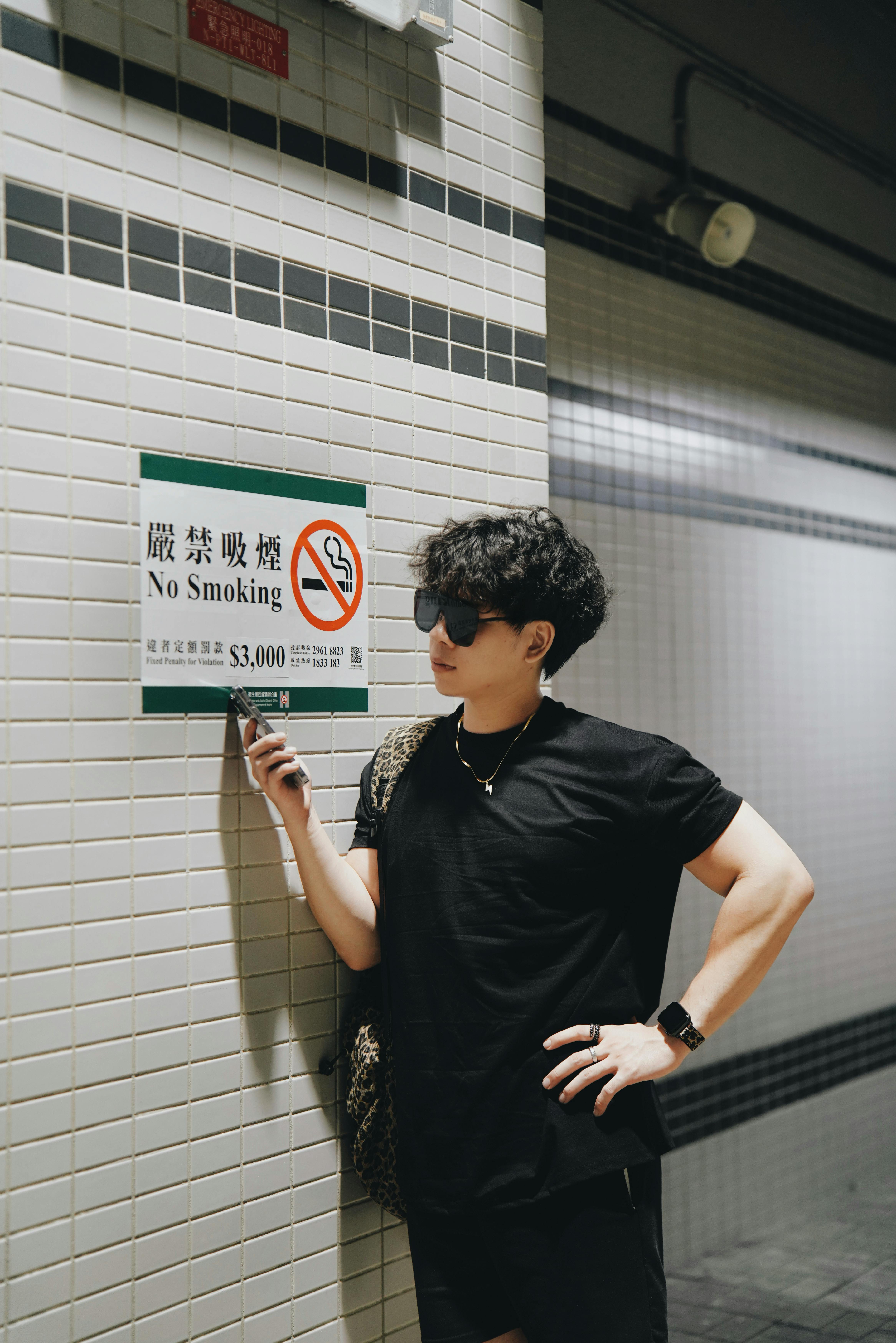 Young man with sunglasses stands near a no smoking sign indoors.