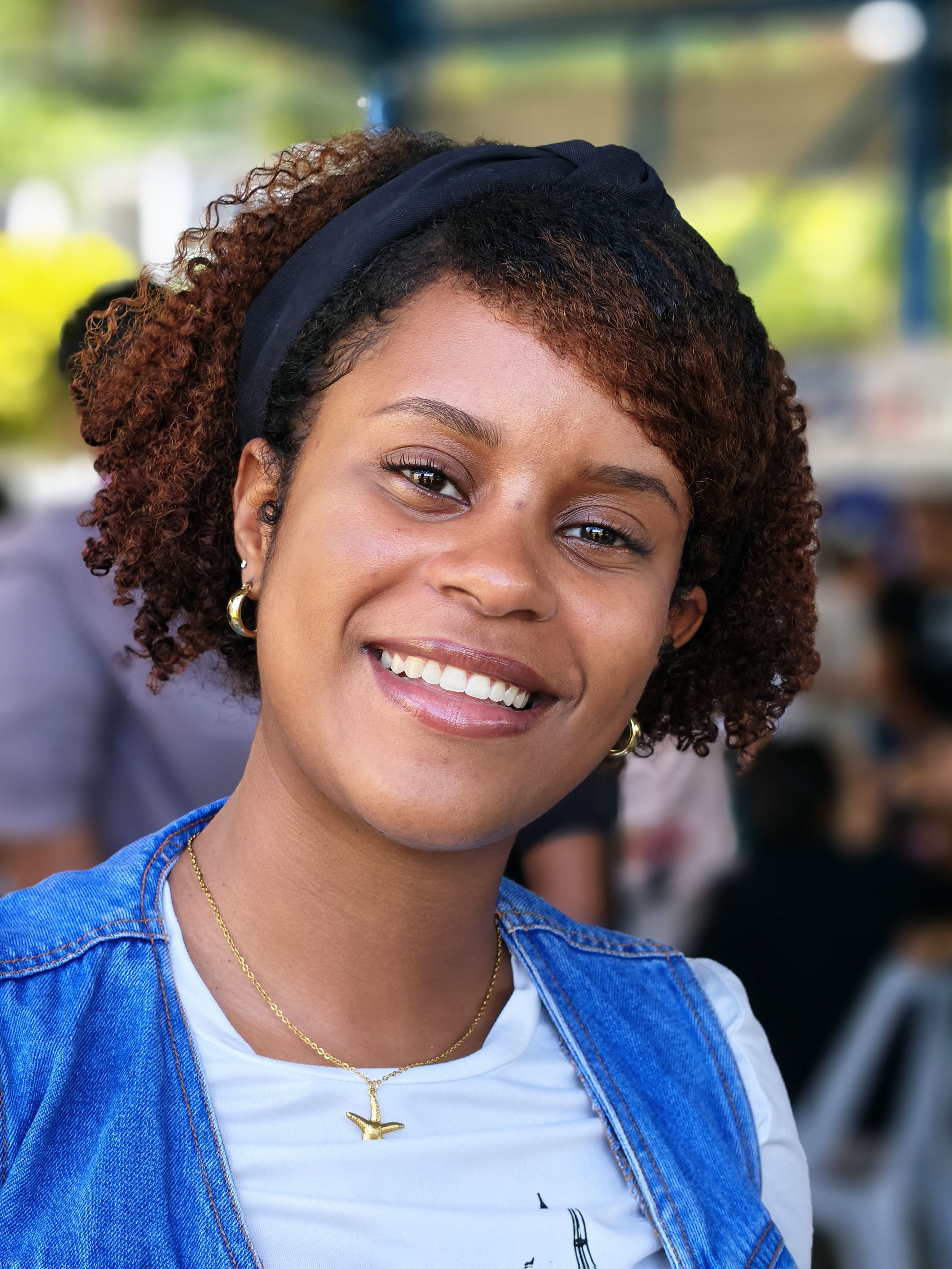 Portrait of a young woman smiling brightly outdoors, showcasing natural curly hair and casual attire.