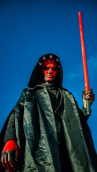 A person in a dramatic sci-fi costume holds a red lightsaber against a clear blue sky.
