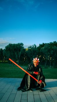 Masked person in dark costume with lightsaber in park under clear sky, Baku.