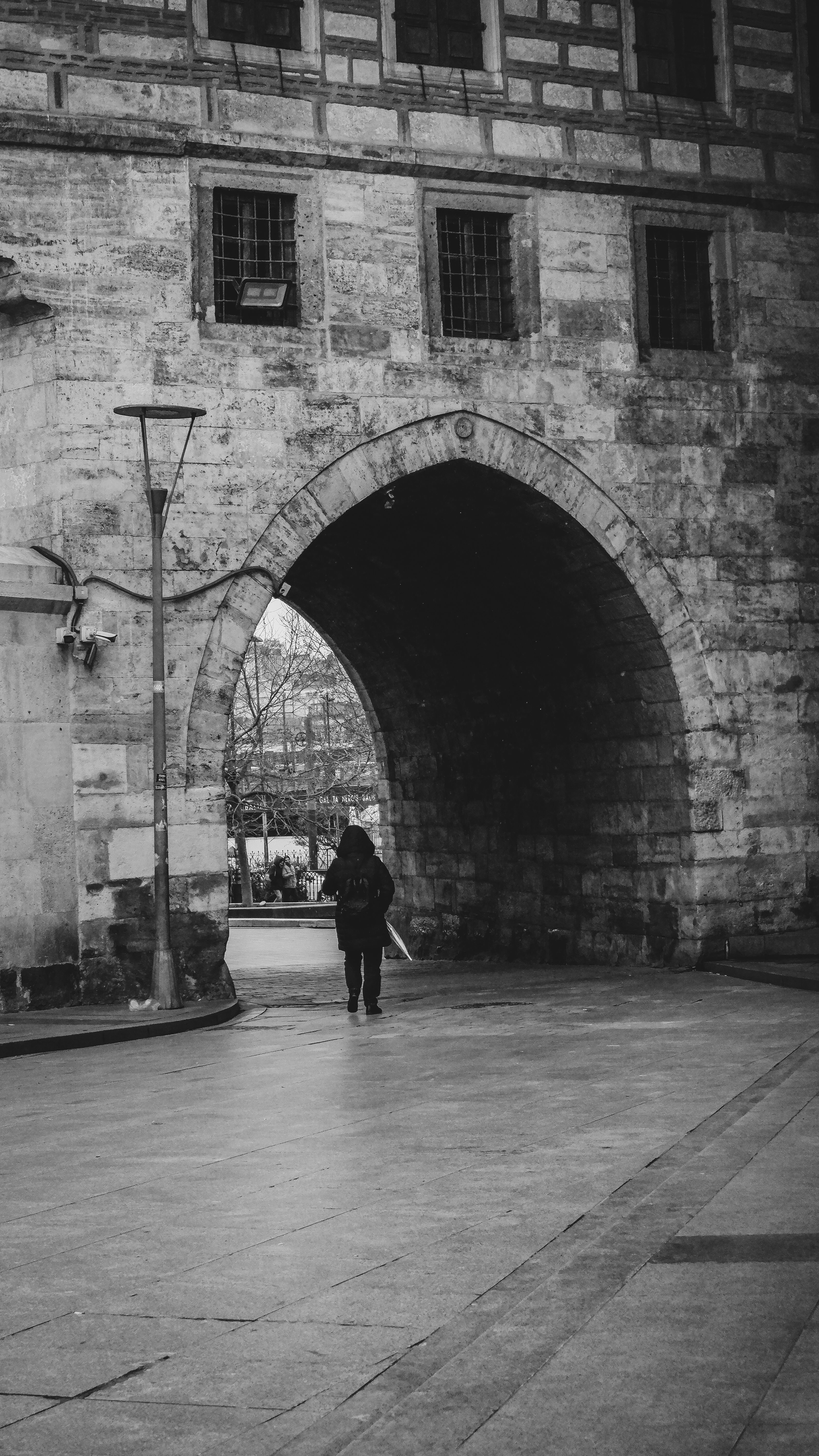 A lone figure walks through a historic stone archway in a black and white cityscape, evoking a sense of solitude and timelessness.