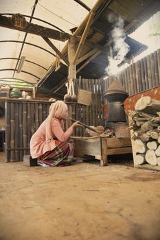 A woman cooks using a traditional stove in a rustic indoor setting with wooden accents and smoke rising.