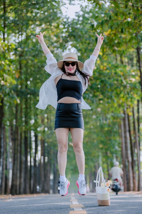 Free Woman wearing summer outfit jumps joyfully on a tree-lined road, showing excitement and freedom. Stock Photo