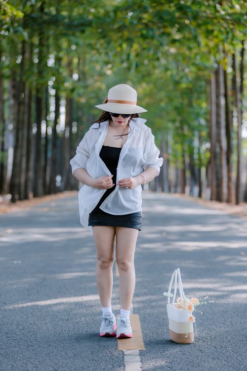 Free Stylish woman in hat and casual summer attire walking along a tree-lined road with a floral tote bag. Stock Photo