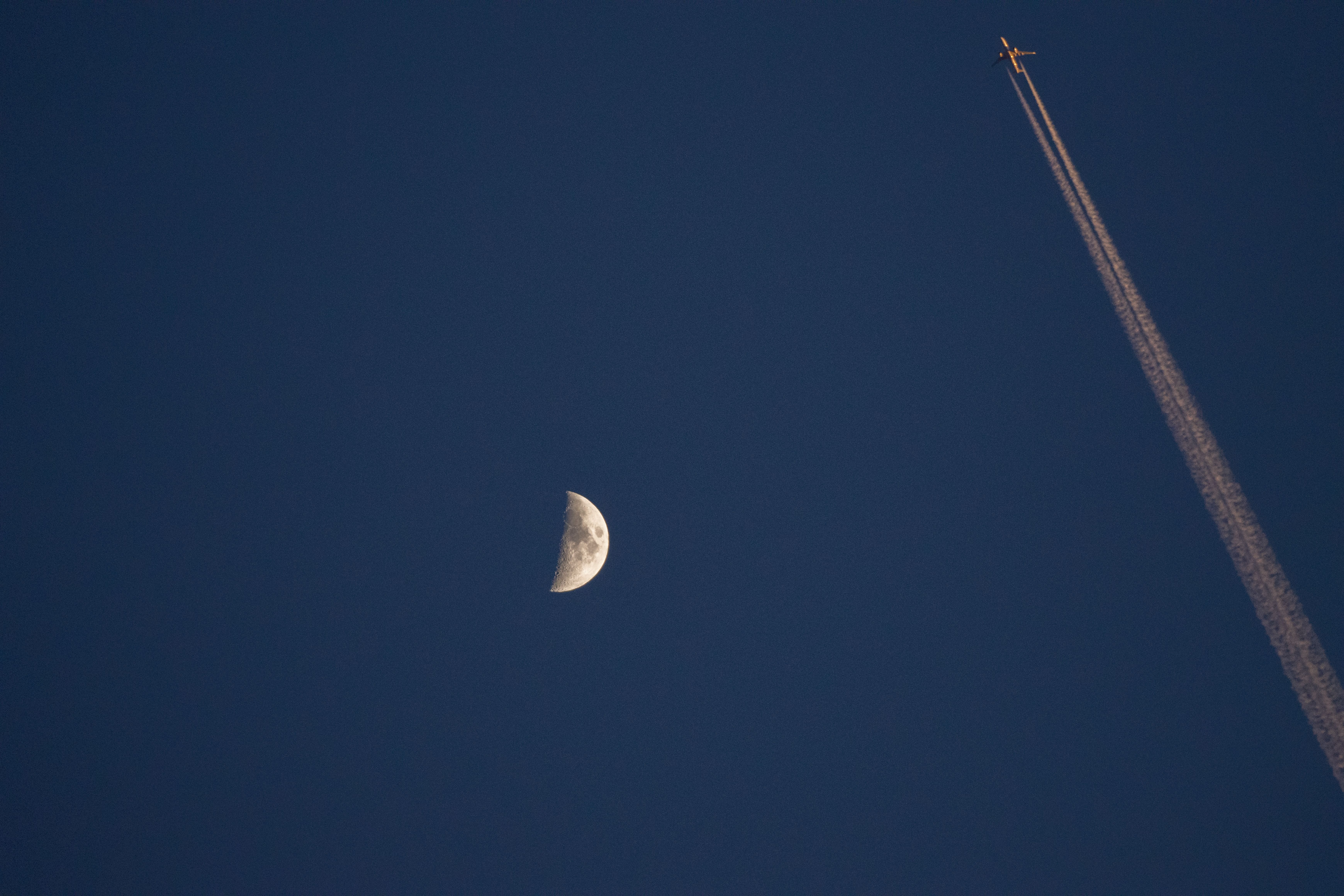 Airplane with contrail passing near the moon against a deep blue night sky, captured over Jönköping.