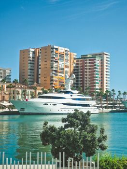 A luxury yacht docked at a marina in Spain with high-rise buildings in the background.