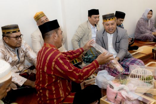 Men in traditional attire exchange gifts indoors, embracing cultural norms.