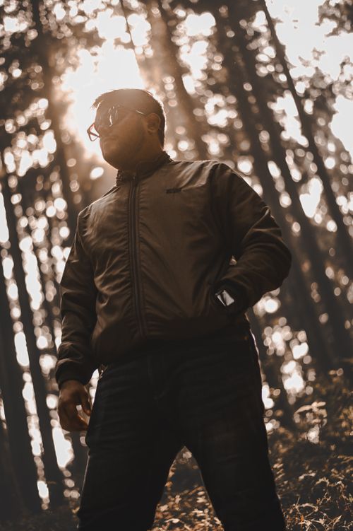 Free A man wearing sunglasses stands in a forest with sunlight filtering through the trees, creating a dramatic scene. Stock Photo