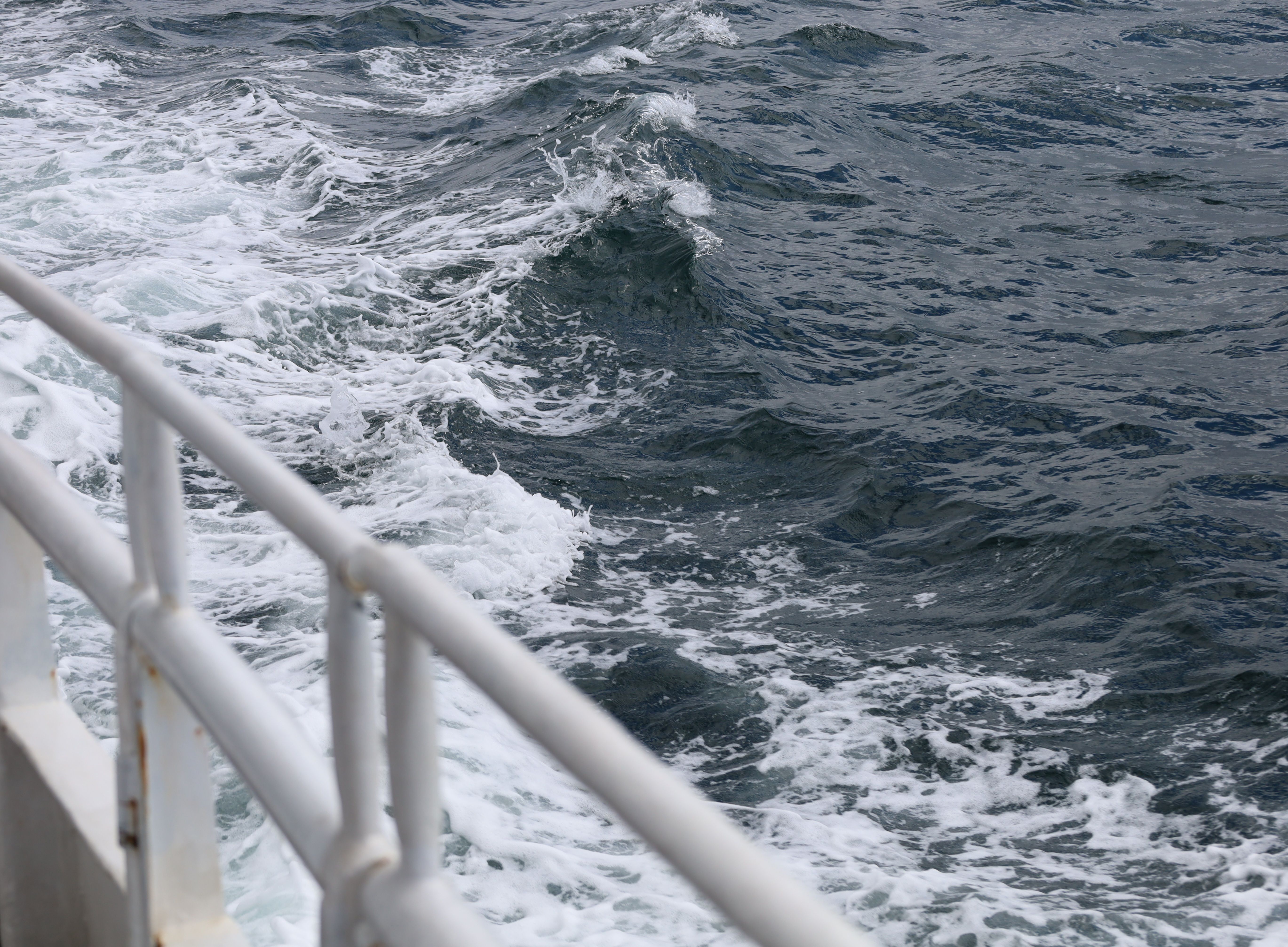 A dynamic view of ocean waves crashing against a ship's railing during the day.