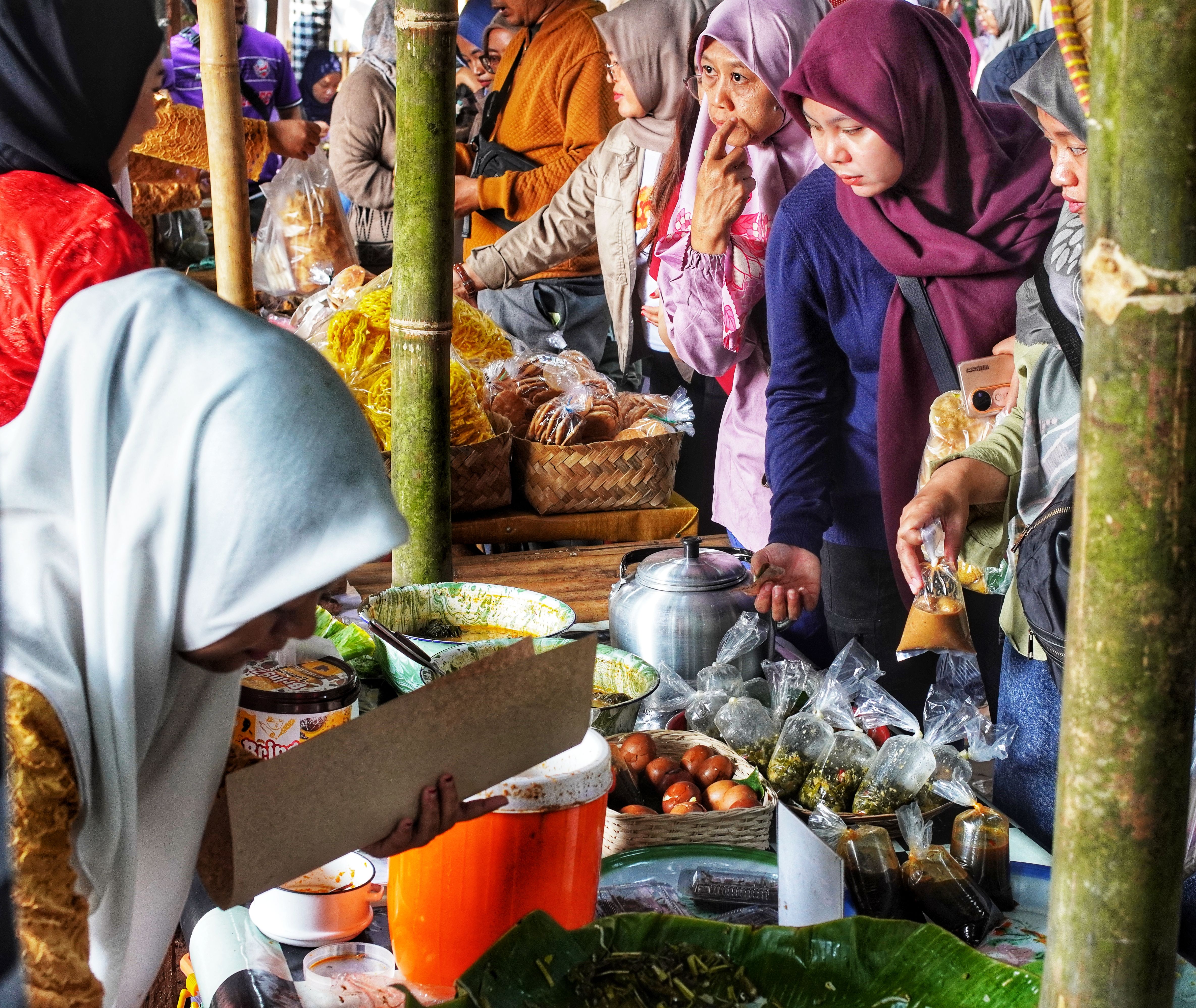 Vibrant women in hijabs interact at a busy Indonesian street market with various goods on display.