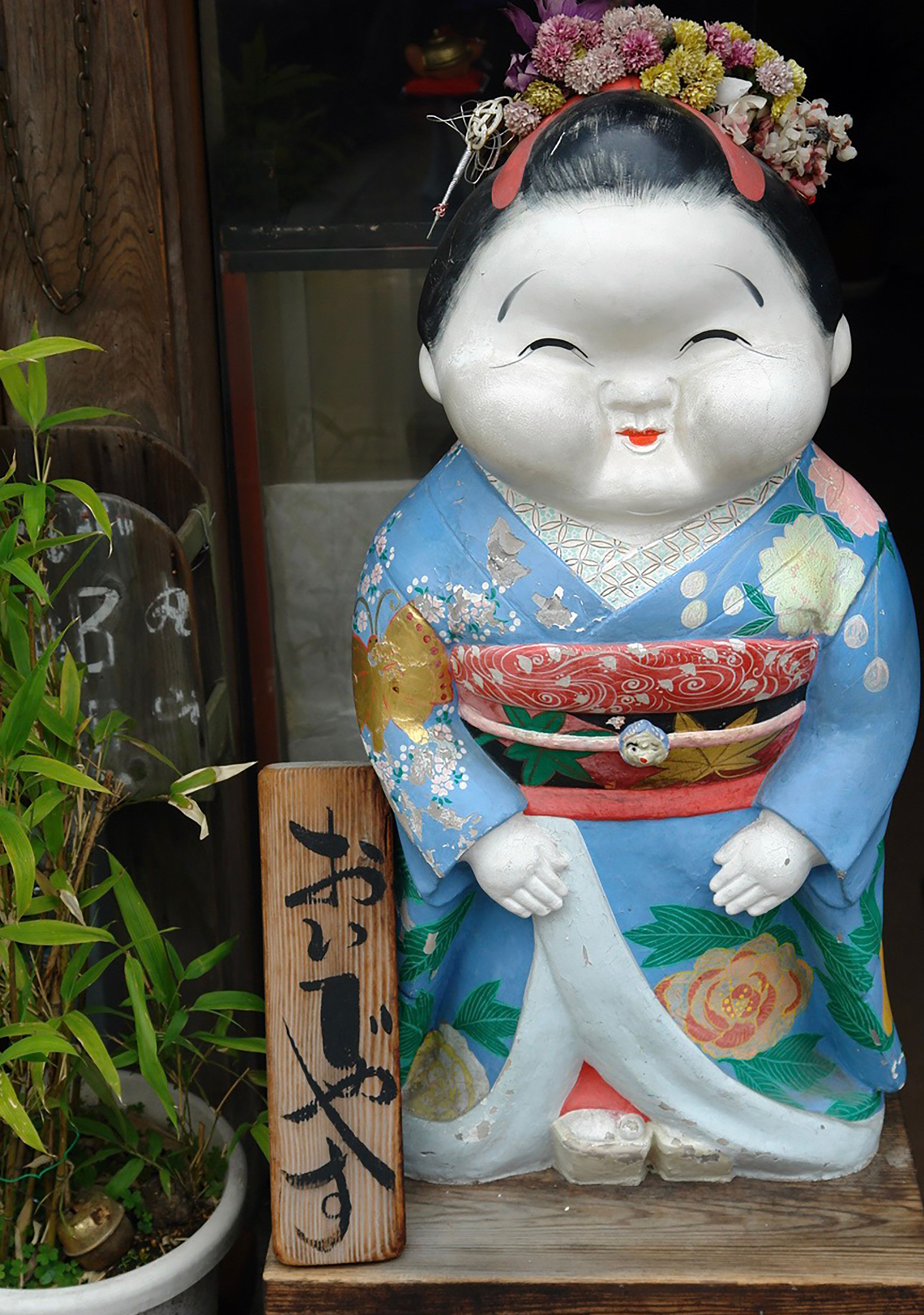A decorative Japanese figurine wearing a vibrant kimono stands beside a wooden sign with Japanese characters.
