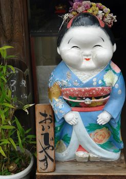 A decorative Japanese figurine wearing a vibrant kimono stands beside a wooden sign with Japanese characters.