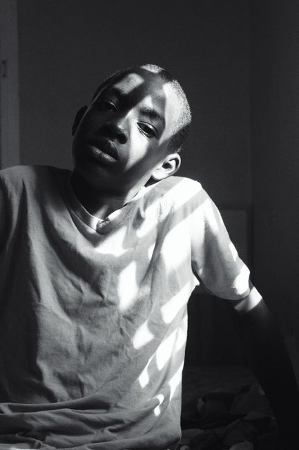 Serious black and white African American boy teenager wearing casual white shirt with shadows on face sitting on bed against window and looking at camera