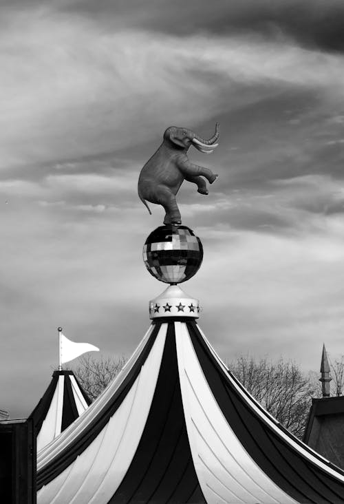 Black and white statue of elephant standing on hind legs on glitter ball placed on circus big top on overcast day