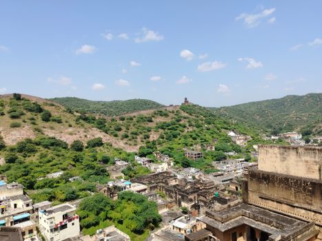A panoramic view of lush green hills surrounding a rustic Indian village under a bright blue sky.