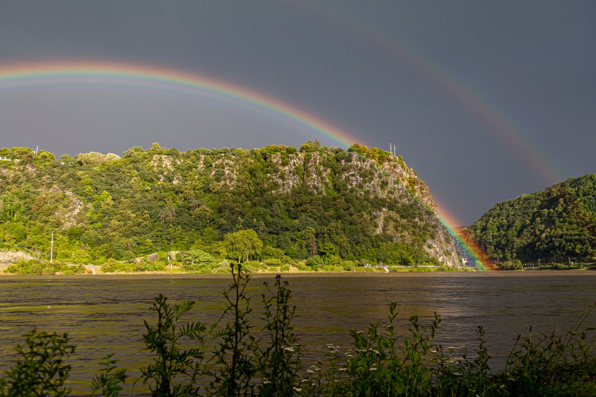 A wooded rock with grey rock faces showing, seen across the river from the water's edge under a stormy sky with a double rainbow