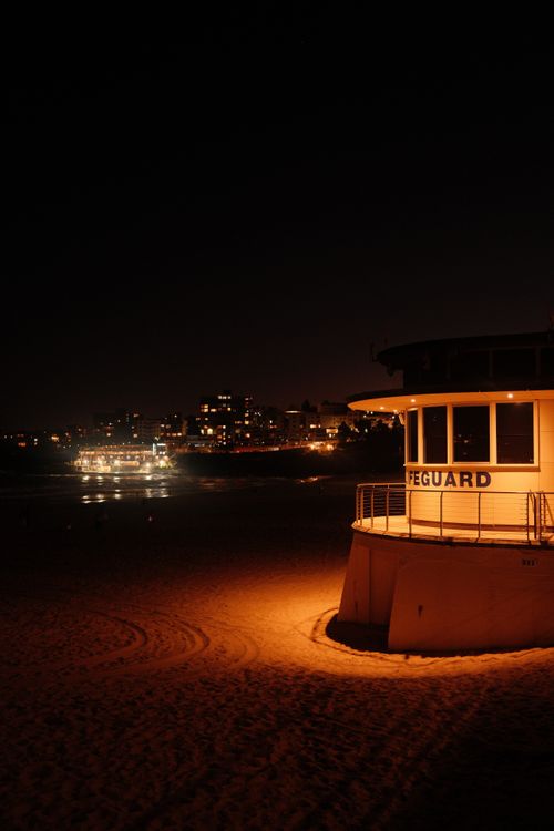 Free A serene nighttime view of Bondi Beach, Sydney, featuring a lifeguard tower illuminated against the city skyline. Stock Photo