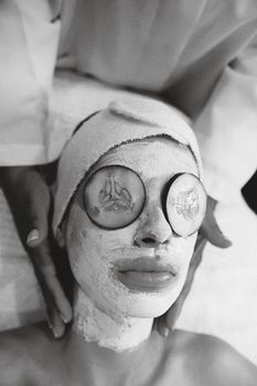 Black and white image of a woman with a face mask and cucumber slices, exuding calmness.
