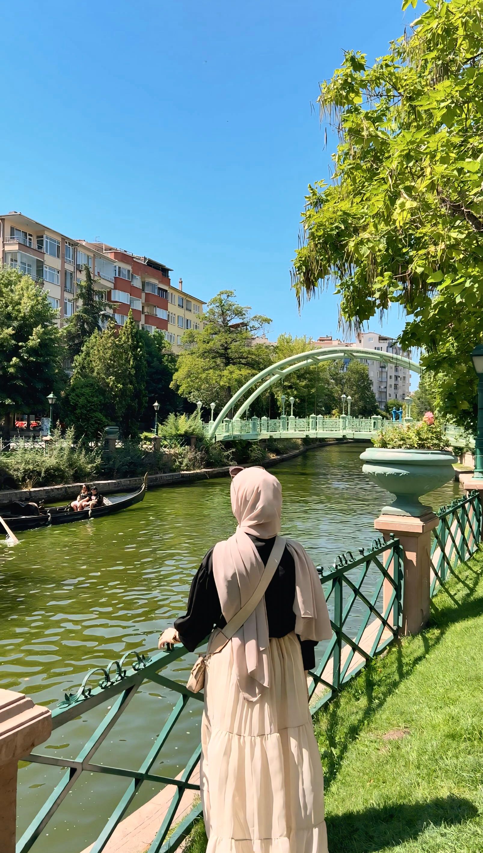 Woman in hijab enjoying a riverside view on a sunny day with a visible bridge and buildings.