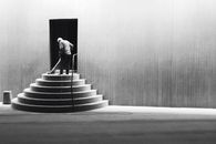 A solitary man sweeps a tiered staircase in a black and white minimalist setting.
