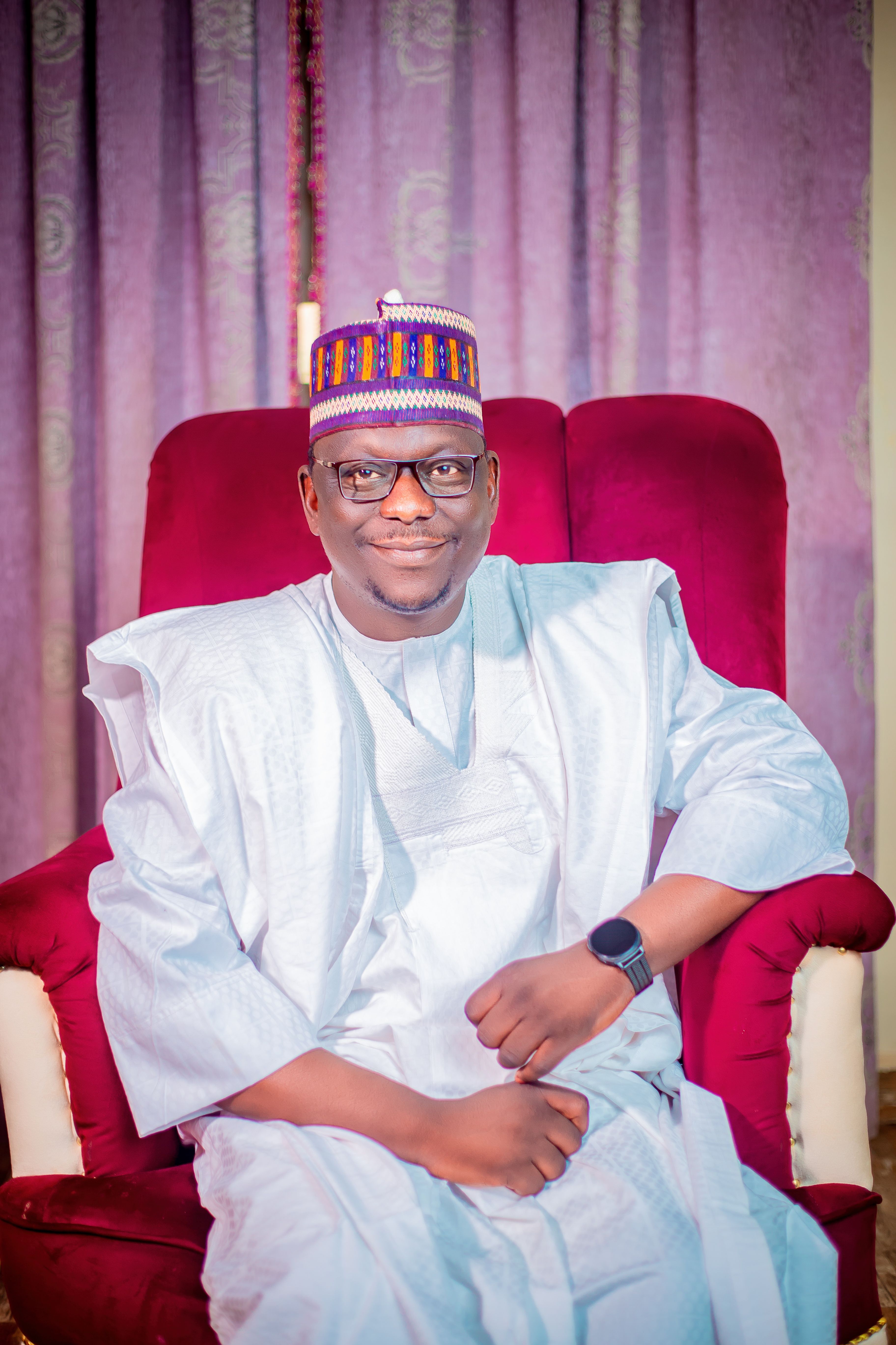 Portrait of a smiling man wearing traditional attire seated on a red chair indoor.