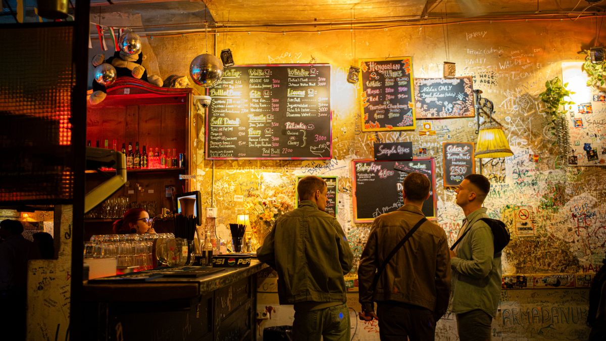 Three people in a Budapest ruin pub, discussing drinks under vibrant lighting and eclectic decor.