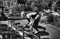 Black and white side view full body sporty swimmer in swimming suit and goggles standing on block in track start position preparing to dive in outside pool