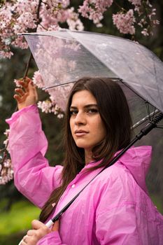 Young woman in a pink raincoat holding an umbrella under blooming cherry blossom branches in a natural outdoor setting.