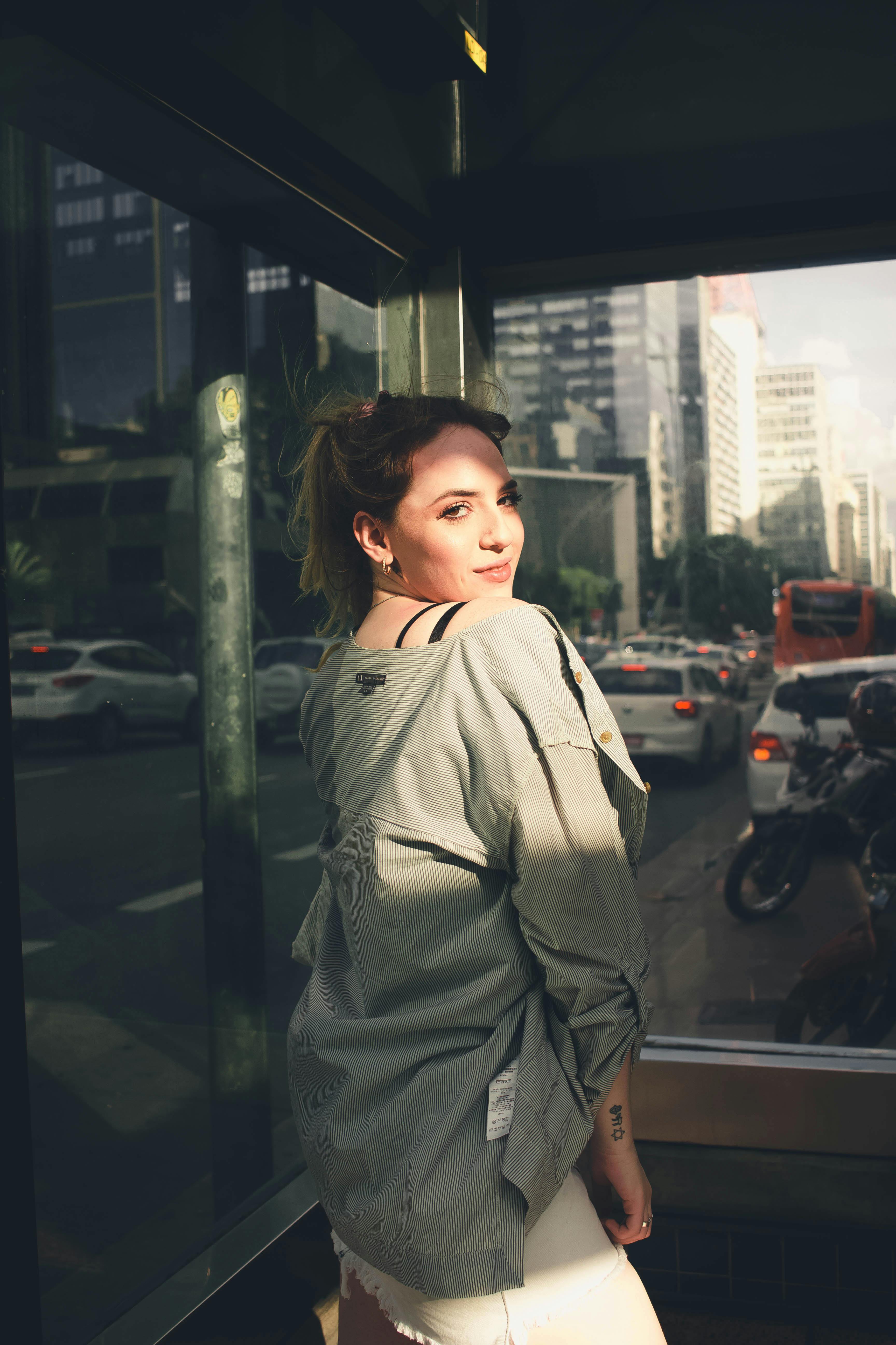 侧面视角下，一位穿着时尚服装的年轻女性背对相机，站在建筑物的玻璃墙前