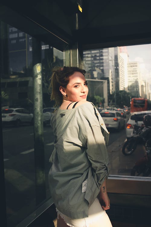 Side view positive content young female wearing stylish outfit standing against glass wall of building and looking at camera over shoulder