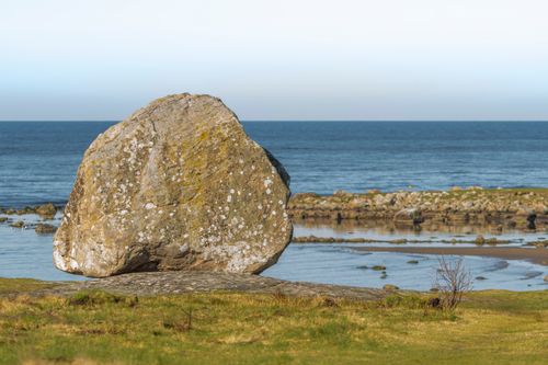Free A large boulder on a grass field overlooking the calm ocean under a clear sky. Stock Photo