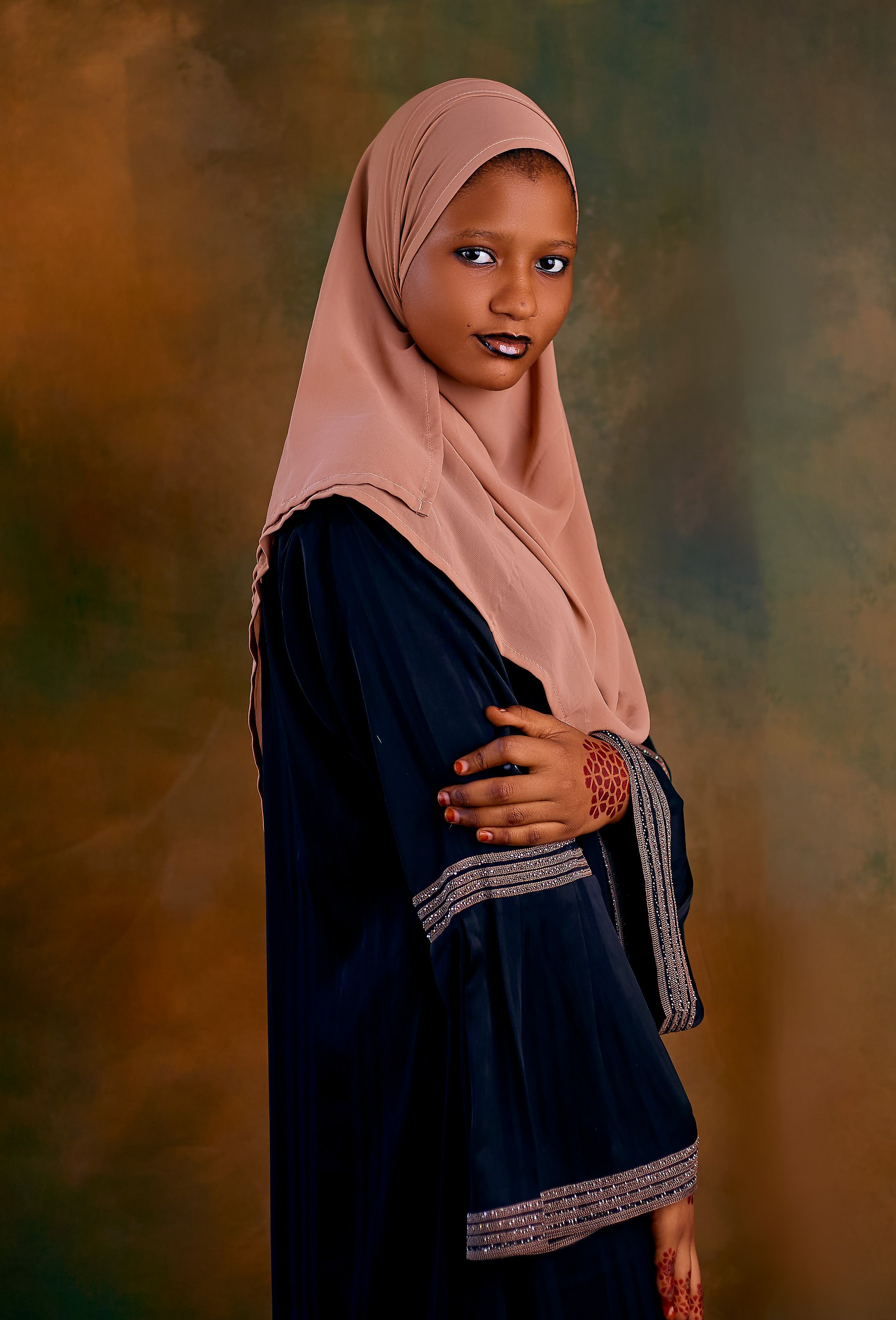 A young woman in a hijab and traditional dress poses against a warm backdrop.