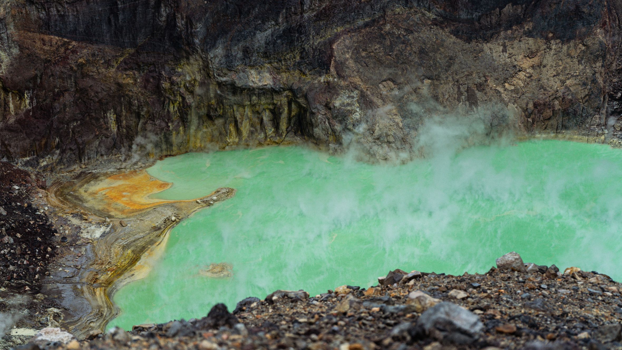 A steaming green acidic lake in a volcanic crater with vibrant color contrast against rocky terrain.